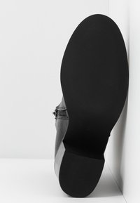 Semelle d'une botte en cuir noire à petit talon montrée sur un fond blanc et un angle de mur.