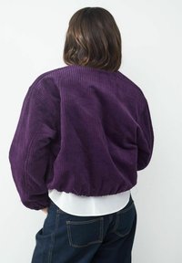 Lila Cordjacke im Bomber-Stil mit geripptem Struktur, gekräuseltem Saum und lässiger Passform, getragen über einem weißen Schichtoberteil und blauen Jeans.