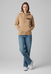 Felpa beige con tasca frontale e testo nero "AMOUR". Indossata con jeans azzurri e sneakers bianche. Il tessuto appare morbido e liscio.