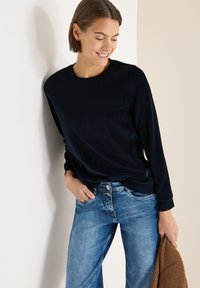 Laivastonsininen pitkähihaisen yläosa pyöreällä pääntiellä, yhdistetty vaaleansinisiin farkkuihin. Yläosassa on sileä tuntuma ja istuvat resorit hihansuissa, jotka on pidetty rentosti vyötäröllä.