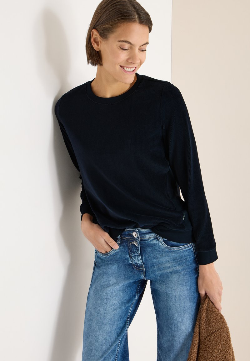 Laivastonsininen pitkähihaisen yläosa pyöreällä pääntiellä, yhdistetty vaaleansinisiin farkkuihin. Yläosassa on sileä tuntuma ja istuvat resorit hihansuissa, jotka on pidetty rentosti vyötäröllä.