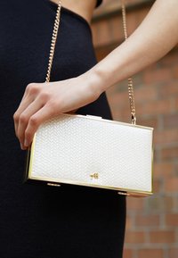 Femme tenant une pochette rectangulaire blanche texturée avec une bandoulière chaîne dorée, portant une robe noire, sur un fond en briques.