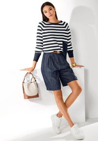 Frau trägt einen navyblauen und weißen gestreiften Langarmshirt, navyblaue Shorts, weiße Sneakers, ein goldenes Armband, hält eine beige und braune Handtasche und lehnt an einer weißen Oberfläche.