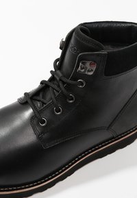 Botín negro de cuero hasta el tobillo con punta redonda, sistema de cordones, ojales de metal y cuello acolchado. Presenta una suela marrón en contraste.