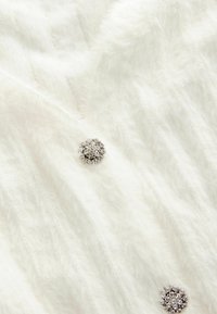 Tissu blanc et moelleux avec une texture en relief ; présente un bouton orné de cristal argenté comme accent décoratif.