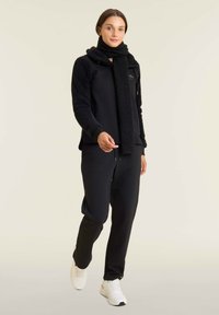 Felpa nera in pile con zip e maniche testurizzate, abbinata a pantaloni jogger neri e a una sciarpa coordinata. Sneakers bianche completano l'outfit.