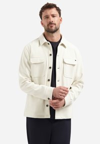 Man in een crème knoopjack met borstzakken over een zwart shirt, die zijn manchetten aanpast en tegen een effen lichte achtergrond staat.