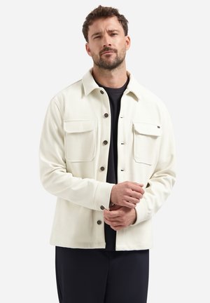 Man in een crème knoopjack met borstzakken over een zwart shirt, die zijn manchetten aanpast en tegen een effen lichte achtergrond staat.