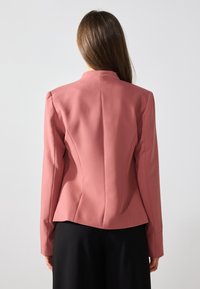 Blazer ajusté en tissu rose doux avec une coupe structurée, épaules rembourrées et coutures discrètes à l'arrière. Associé à un pantalon noir.