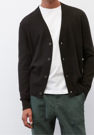 Uomo che indossa un cardigan nero con bottoni sopra una t-shirt bianca e pantaloni verde scuro, in piedi contro uno sfondo chiaro e semplice.