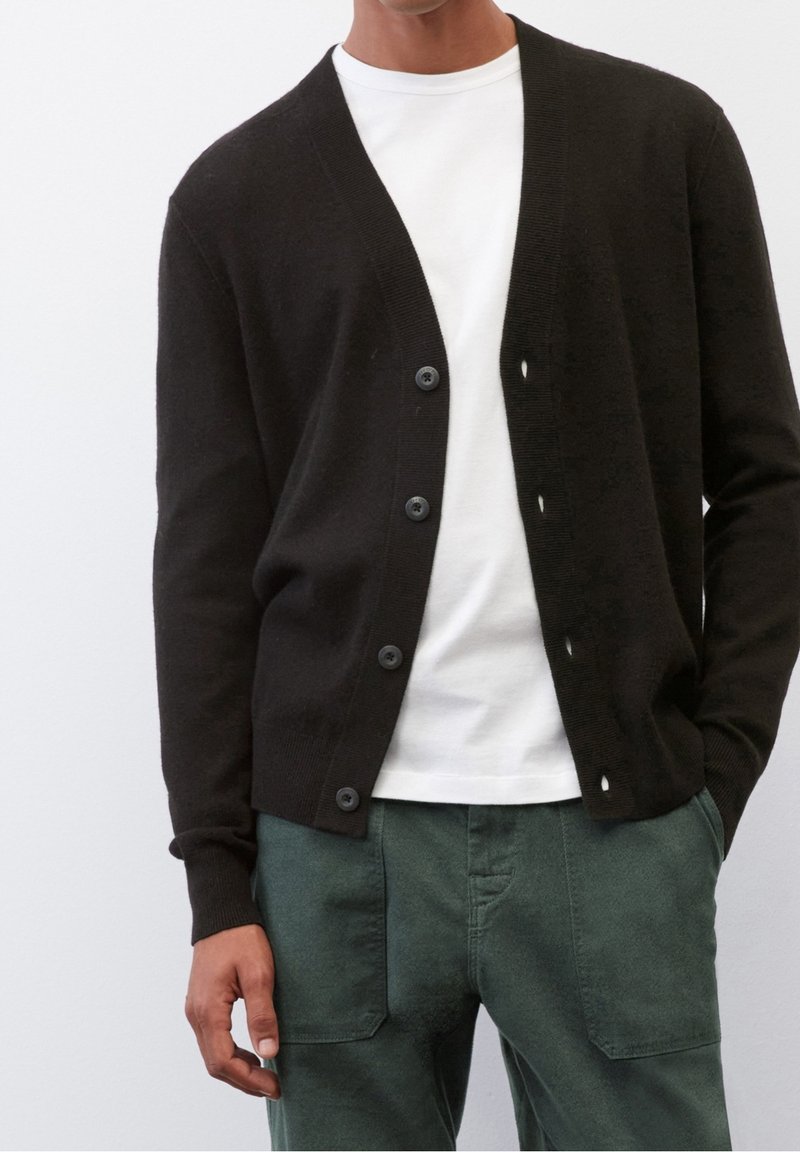 En man som bär en svart knappad cardigan över en vit t-shirt med mörkgröna byxor, står mot en ljusbakgrund.