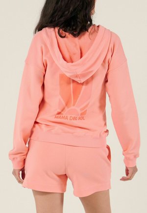 Vrouw draagt een koraalroze hoodie en korte broek, op de achterkant van de hoodie staat een zonafbeelding met de tekst "Mama Del Sol".