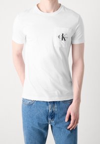 Muž v jednoduchém bílém tričku s krátkým rukávem a náprsní kapsou, na které je logo "CK Calvin Klein Jeans", v kombinaci s modrými džíny.