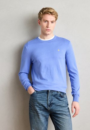 Light blue knit sweater with long sleeves and a round neckline, featuring a small gold emblem on the left chest. Paired with faded blue jeans.