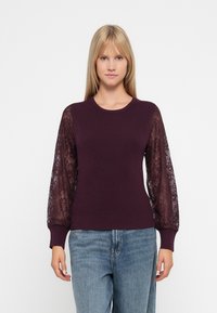 Burgundy pulover s rebrastim tijelom, s prozirnim čipkastim rukavima i rebrastim manšettama. Uparen s svijetloplavim trapericama.