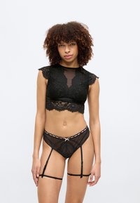 Soutien-gorge bralette en dentelle noire avec manches courtes, motif floral et bords festonnés, assorti à une culotte transparente noire à pois avec bretelles porte-jarretelles.