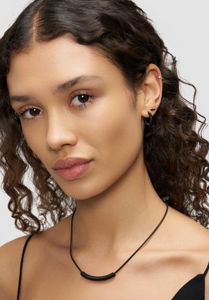 Schwarze Halskette mit einem gebogenen Metallakzent an einem dünnen Band. Modell mit lockigem Haar trägt kleine Creolen und dezentes Make-up. Neutrale Hintergrundfarbe.