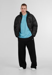 Giacca a vento nera con motivo testurizzato, t-shirt azzurra chiara e pantaloni della tuta neri. Sneakers bianche completano il look informale.