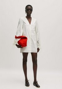 Weißes Hemdkleid mit langen Ärmeln und Spitzenbesatz. Das Model hält eine rote Tasche, trägt schwarze Sonnenbrillen und schwarze Schuhe. Einfaches, minimalistisches Design.