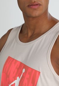 Beige linne i bomull med ett stort rött grafiskt tryck med texten "HIGH FLY" och en silhuett av en basketboll. Slät textur.