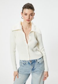 Cardigan bianco a costine con colletto in pelliccia sintetica, dotato di chiusura frontale a bottoni e vestibilità slim, abbinato a jeans azzurri a vita alta.