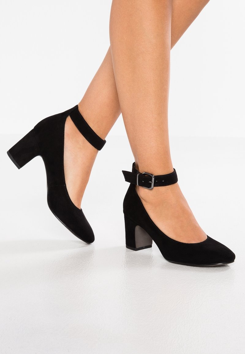 Talons carrés en daim noir avec bride à la cheville, portés pieds nus, talon de hauteur moyenne et fermeture à boucle argentée sur la sangle.