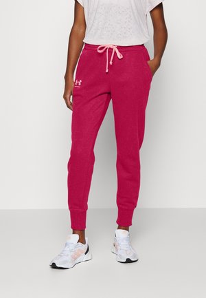 Person iført pink Under Armour joggers med snor i taljen og hvide sneakers, stående med den ene hånd i lommen.