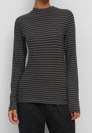 Persoon die een strakzittend, zwart en donkergrijs horizontaal gestreept shirt met lange mouwen draagt, en donkere gestructureerde broek, staand tegen een effen achtergrond.