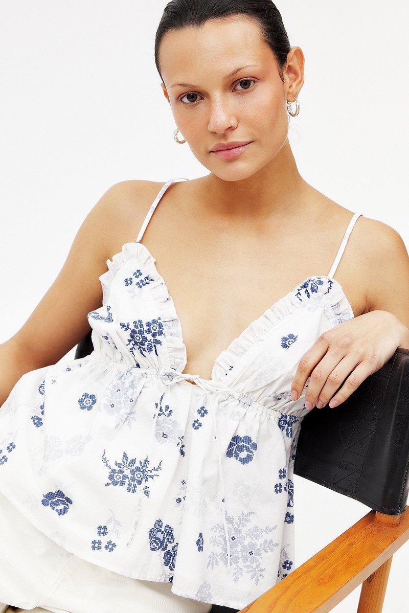 Floral blouse with ruffled neckline, white fabric featuring blue patterns. Adjustable straps, loose fit, and gathered waist detail.