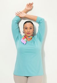 Camicia turchese a maniche lunghe con scollo a V, abbinata a una sciarpa a righe multicolori attorno al collo. Tessuto morbido, vestibilità rilassata.