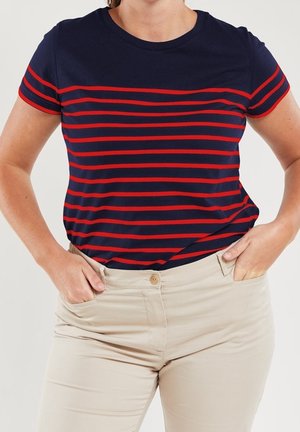 Vrouw draagt een donkerblauw t-shirt met rode horizontale strepen en beige broek, staat met handen in de zakken tegen een effen achtergrond.