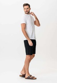 Mand i hvid t-shirt, sorte shorts og brune sandaler står barfodet og kigger over sin skulder mod en ensfarvet hvid baggrund.