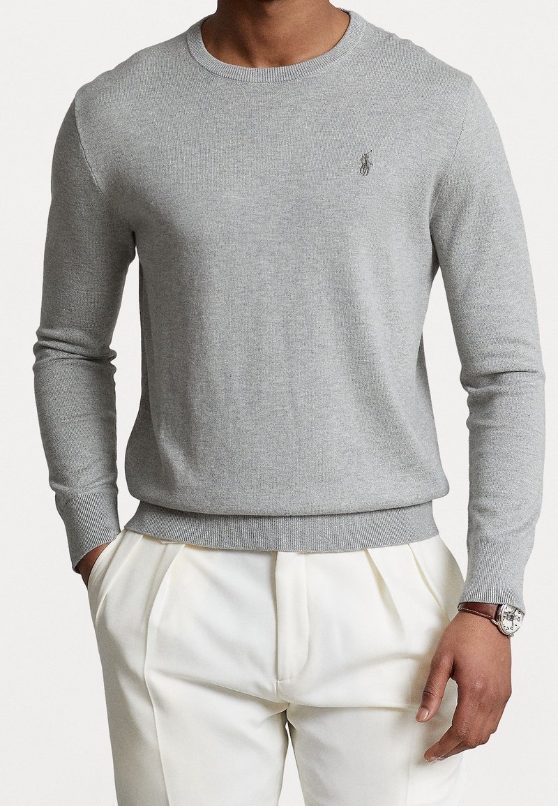 Homme portant un pull gris clair à manches longues avec un petit logo sur la poitrine, un pantalon blanc plissé, et une montre-bracelet avec un bracelet en cuir marron.