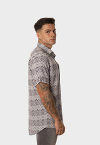 Camisa de manga corta en gris con un patrón en zigzag. Presenta un cuello de botón y un corte relajado. La tela tiene una textura suave.