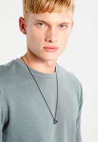 Sort kædannecklace med en trekantet vedhæng båret over en lys grøn langærmet skjorte. Vedhænget har et minimalistisk design.
