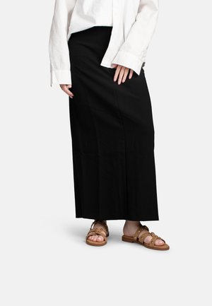 Persoon die een lange zwarte rok, een oversized wit overhemd en beige studs vlakke sandalen draagt, staand tegen een effen achtergrond.
