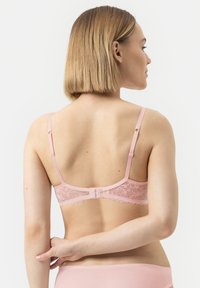 Vrouw met kort blond haar die een lichtroze kanten beha van achteren vastmaakt, met bijpassend roze ondergoed tegen een eenvoudige witte achtergrond.