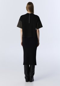 Donna con capelli lunghi indossa un top corto nero con zip sul retro, una gonna midi a coste e stivali neri fino al ginocchio, in piedi su uno sfondo neutro.