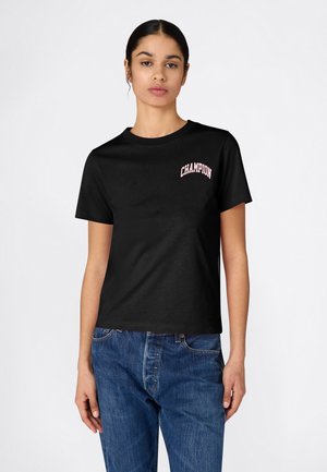 Frau trägt ein schwarzes kurzärmliges T-Shirt mit dem Schriftzug "CHAMPION" auf der Brust, kombiniert mit blauen Jeans, und steht vor einem schlichten Hintergrund.