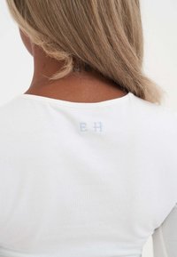 Ein blonder Mensch mit mittlerem Hautton trägt ein weißes Oberteil, das den Rückenausschnitt zeigt, mit kleinen bestickten Buchstaben "E H" in hellblau.