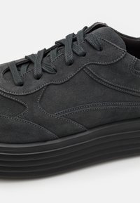 Gros plan d'une basket en daim noir avec un design à panneaux cousus, des lacets noirs et une semelle en caoutchouc épaisse et texturée.