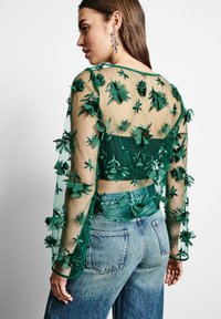 Grüner transparenter Bluse mit floralen Stickereien, langen Ärmeln und einem verkürzten Schnitt, kombiniert mit hellblauen High-Waist-Jeans.
