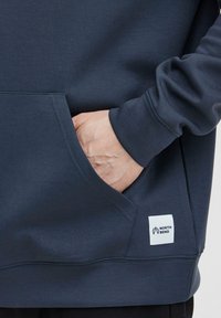 Tumši zils krekls ar kangaroo kabatu, ribotiem piedurknēm un baltu etiķeti ar uzrakstu "NORTH BEND" tuvāk apakšai. Kokvilnas maisījuma materiāls.