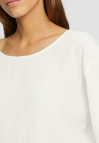 Haut texturé blanc avec un large décolleté et des manches trois-quarts, présentant un subtil motif en relief sur le tissu.