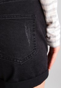 Black denim shorts with a back pocket. Features a frayed detail and rolled hem. Fabric appears smooth with a slight sheen.