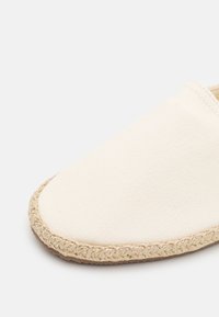 Kangasliukusandaali vaaleassa värissä, jossa on teksturoitu päällinen ja punottu juuttipohja. Pyöreä varvasosa, jossa on ompeluyksityiskohtia reunoilla.
