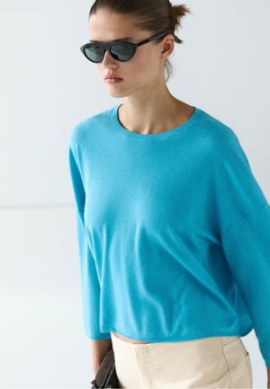 Kvinde iført sorte solbriller og en klarblå bluse med lange ærmer samt beige bukser mod en ensfarvet lys baggrund.