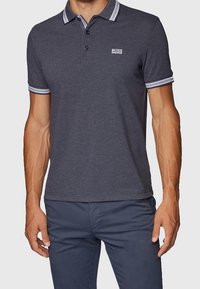 Mörkgrå polo med två knappar, vitrandig krage och liten logotyp på bröstet. Bärs med mörkfärgade byxor.