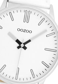 Montre argentée avec un cadran blanc, aiguilles des heures et des minutes noires, marqueurs d'heures minimalistes et un bracelet en cuir blanc. Nom de la marque : OOZOO.