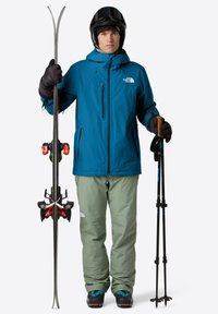 Uomo con giacca blu, pantaloni verdi, casco nero e guanti, che tiene gli sci in posizione verticale in una mano e i bastoncini da sci nell'altra, indossando scarponi da sci.